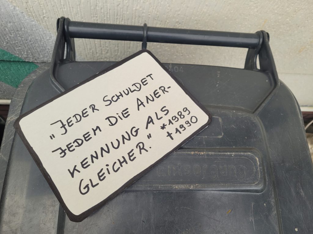 Schild mit Zitat "Jeder schuldet jedem die Anerkennung als Gleicher." auf Mülltonne